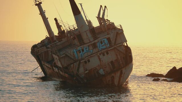 A rusting shipwreck stands majestically against a glowing sunset backdrop, hull and cranes silhouetted by the warm evening hues.
