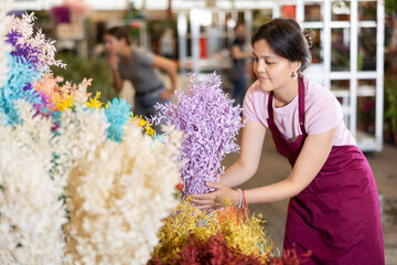 Smiling asian young female florist working at flower market, placing bunches of colorful dried...