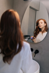 Vertical shot of young woman playing with long red hair looking in mirror standing in bathroom in morning. Reflection on mirror of pretty redhead female preens, admires, straightening hairstyle