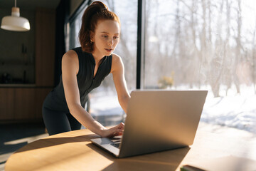 Focused sporty young woman in outfit using laptop standing at desk by window on sunny day, thinking...