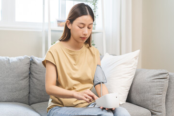 Hypotension problem asian young woman sitting on couch checking high blood pressure and heart rate...