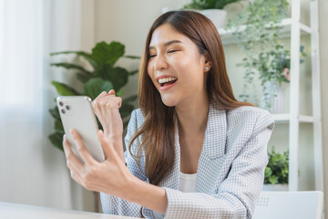 Happy cheerful asian young woman celebrate online looking at smart phone on mobile app screen,...