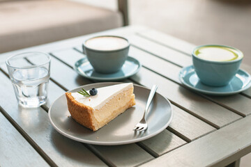 A piece of classic cheesecake decorated with a berry and rosemary on a plate, hot drinks on the table on the terrace.