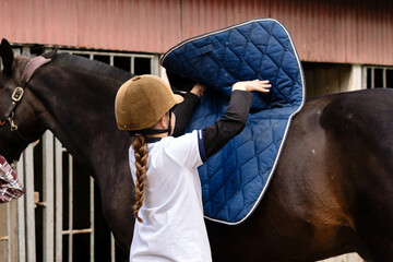 Girl placing saddle pad on horse's back at stable.