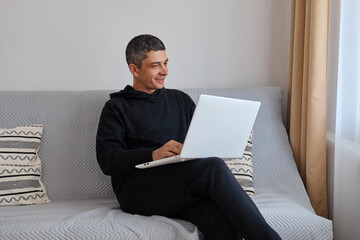 Young attractive smiling guy wearing black clothing browsing at his laptop sitting at home on cozy sofa at home doing his remote work looking smiling at computer display