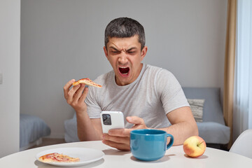 Angry mad Caucasian man wearing T-shirt sitting at table eating slice of pizza screaming with aggressive expression using smartphone receives massages with bad news in home interior