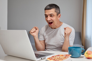 Overjoyed extremely happy Caucasian man wearing gray T-shirt working on laptop screaming exclaiming with clenched fists celebrating his success in home interior