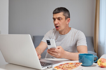 Shocked Caucasian man wearing gray T-shirt working on laptop and using his smartphone expressing astonishment looking at computer display with open mouth in home interior