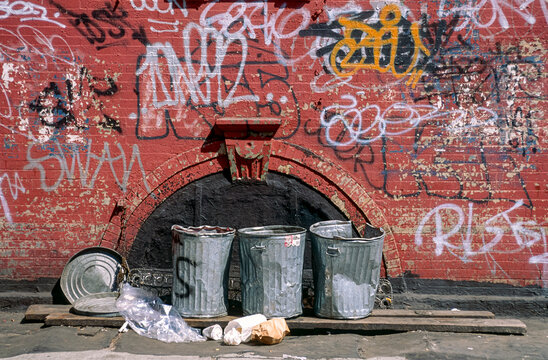 Eingescanntes Diapositiv einer historischen Farbaufnahme von verbeulten M&uuml;lleimern mit verstreuten M&uuml;llt&uuml;ten vor einer Hauswand mit Graffities, New York, Harlem, USA