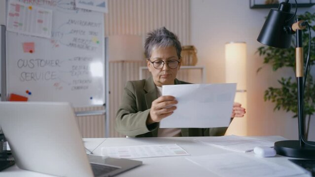 Tired mature businesswoman looks at company statistics documents, work overload