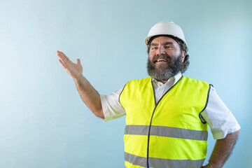 smiling adult man engineer with beard and wearing helmet and safety vest looking at camera on blue background, pointing hands to the left