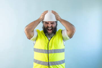 Naklejka premium stressed adult man engineer with beard and wearing helmet and safety vest, looking at camera on blue background holding his head