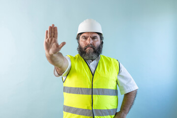 serious adult man engineer with beard and wearing helmet and safety vest, looking at camera on blue...