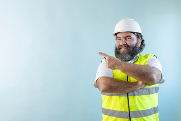 smiling adult man engineer with beard and wearing helmet and safety vest, looking at camera on blue...