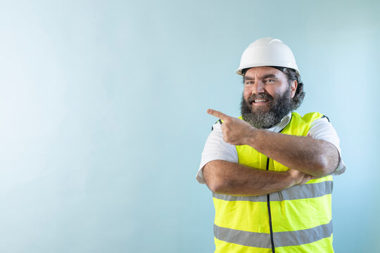 smiling adult man engineer with beard and wearing helmet and safety vest, looking at camera on blue background, with crossing arms