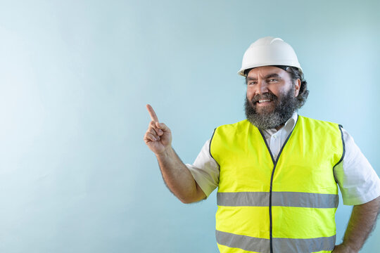 smiling adult man engineer with beard and wearing helmet and safety vest, looking at camera on blue background and pointing finger to the left