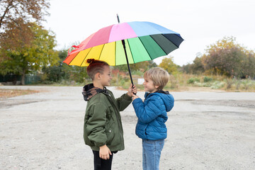 two children, boys 7 years old, stand together under a large rainbow umbrella. autumn atmosphere. symbol of the rainy season, wet autumn weather. Friendship, mutual support
