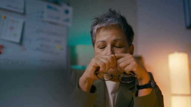 Exhausted woman in her 50s taking glasses off, working in office till late night