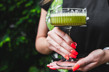 Vibrant green juice in a glass held by hands with red nail polish against a blurred greenery background