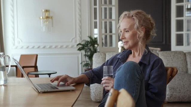 Senior woman sitting at table in living room with laptop. She works remotely or enjoys leisure activities online shopping typing on keyboard. Satisfaction and fulfillment in the older woman life.