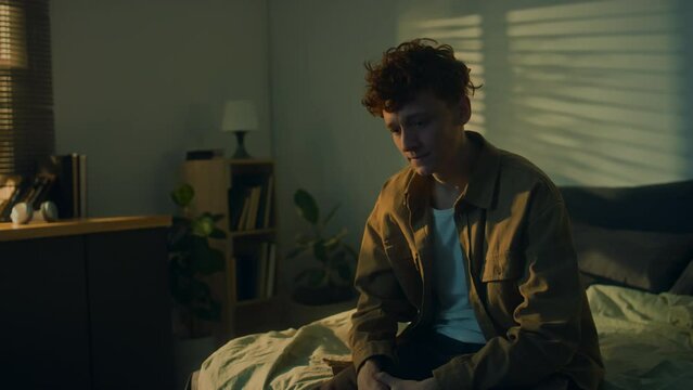 Side handheld footage of Caucasian man suffering from anxiety or despair sitting alone on bed at home