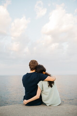 Couple sitting together on a pier, embracing and looking out over the calm sea under a cloudy sky,...