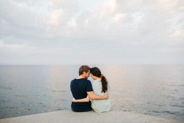 Couple sitting together on a pier, embracing and looking out over the calm sea under a cloudy sky,...