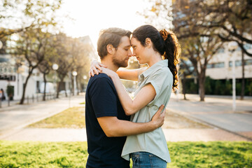 Fototapeta premium Young couple embracing, foreheads touching, in an intimate moment outdoors with trees and buildings in the background.