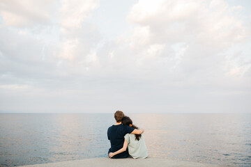 Couple sitting together on a pier, embracing and looking out over the calm sea under a cloudy sky,...