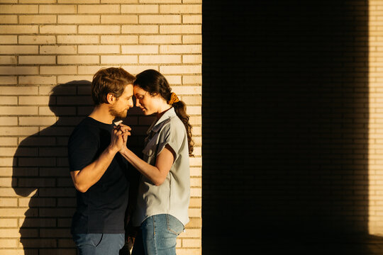 Couple sharing an intimate moment, standing close and holding hands, while the golden light of the setting sun illuminates a brick wall.