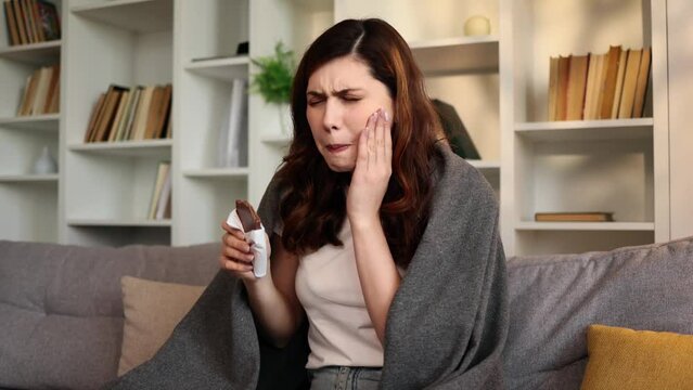Portrait of young brunette woman eating chocolate bar and having toothache sitting on couch at home Sad female feeling pain in tooth because caries gum disease or sensitive teeth indoors