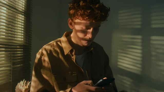 Chest up footage of worried Caucasian Gen Z guy receiving message on mobile at home and getting into panic cautiously looking through blinds outside