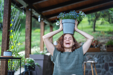 Young happy funny Caucasian woman holding pot of flowers above her head while sitting on wooden terrace of country cottage, summer lifestyle portrait