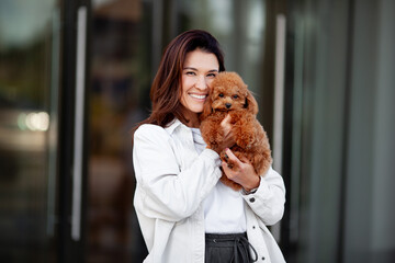 Beautiful happy dark haired woman holds and hugs small dog poodle, looking at camera and smiling against gray background. Moment of joy and affection between the woman and her pet outdoors