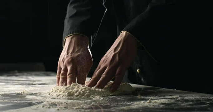 Super slow motion close up of professional artisan baker chef kneads dough with flour while making homemade bread, pasta or pizza on rustic wooden table in traditional bakery kitchen.