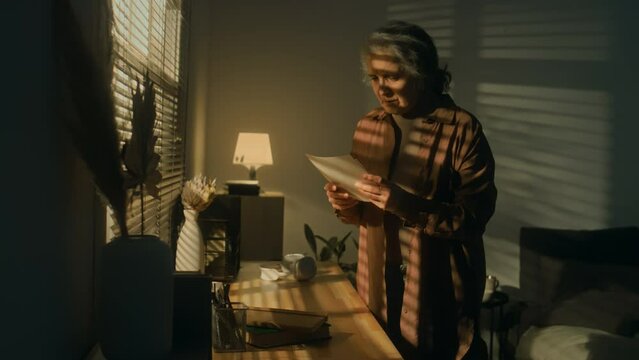 Side medium footage of senior Caucasian woman reading obituary notice or letter with grieving news and feeling worried and anxious while standing by window in light of rising sun rays
