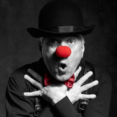 Dramatic selective color portrait of a surprised clown against a dark background with hands on chest.