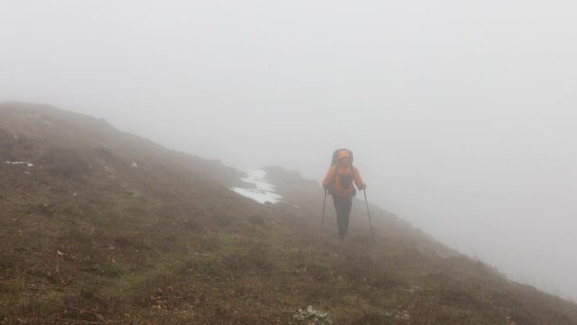 A lone hiker trekking through snowy patches in a mountainous landscape under a cloudy sky during winter.