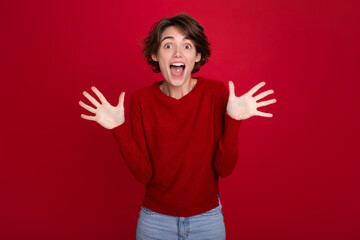 Excited happy funny cute girl with a cool short haircut in a red sweater shows her palms and shouts isolated on a red bright vivid background. Win, lottery, bet, sale, graduation, good news