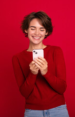 Naklejka premium Vertical photo of a smiling beautiful young modern woman who using a smartphone isolated on a red bright wall. Shopping, chatting, surfing the internet, betting, social media, win in games