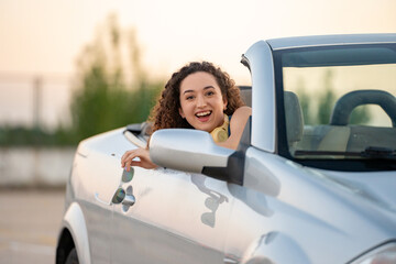 Cheerful young woman with curly hair and headphones enjoys a sunset ride in a convertible car. 