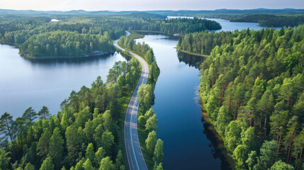 Obraz premium Aerial view of a winding road cutting through lush green woods, flanked by serene blue lakes under the summer sun in Finland, highlighting the natural beauty.