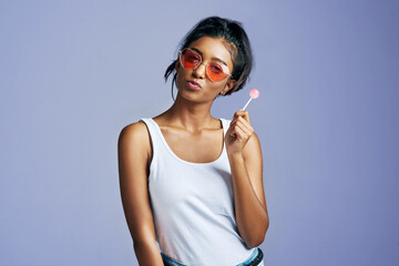 Girl, lollipop and confident with sunglasses in studio on purple background in casual, style and elegant. Portrait, fashion and happy or smile for eyewear or street wear, outfit and satisfied