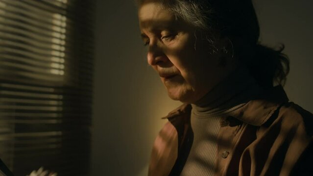 Side closeup of senior Caucasian lady reading obituary notice or letter with frustrating news feeling grief and looking broken while standing by window in dark room