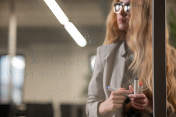Caucasian woman with glasses writes text in English on a glass wall. 