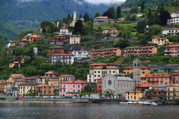 Fototapeta premium A landscape with a view of Lake Como and the Chiesa Santissima Trinita church in the town of Argegno with the mountains in the background covered with greenery and partly with clouds