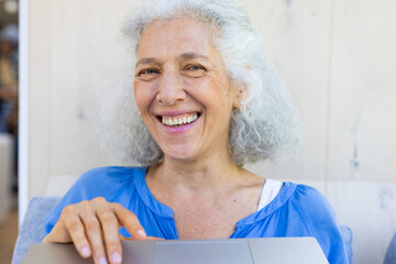 Caucasian senior woman in blue shirt smiles at laptop during home video call