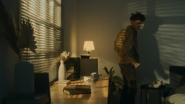 Side handheld footage of young Caucasian man looking at photo at night and suddenly getting mad throwing stuff from table and shouting at home