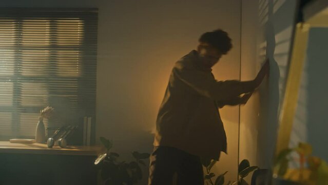 Side handheld slowmo of young curly haired man punching wall in rage and walking away in dimly lit room at dawn