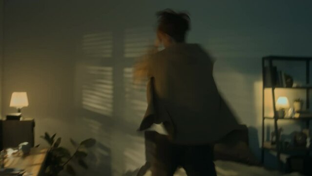 Handheld slowmo of young Caucasian man in rage walking around room and throwing things on floor while going through anger attack alone at night